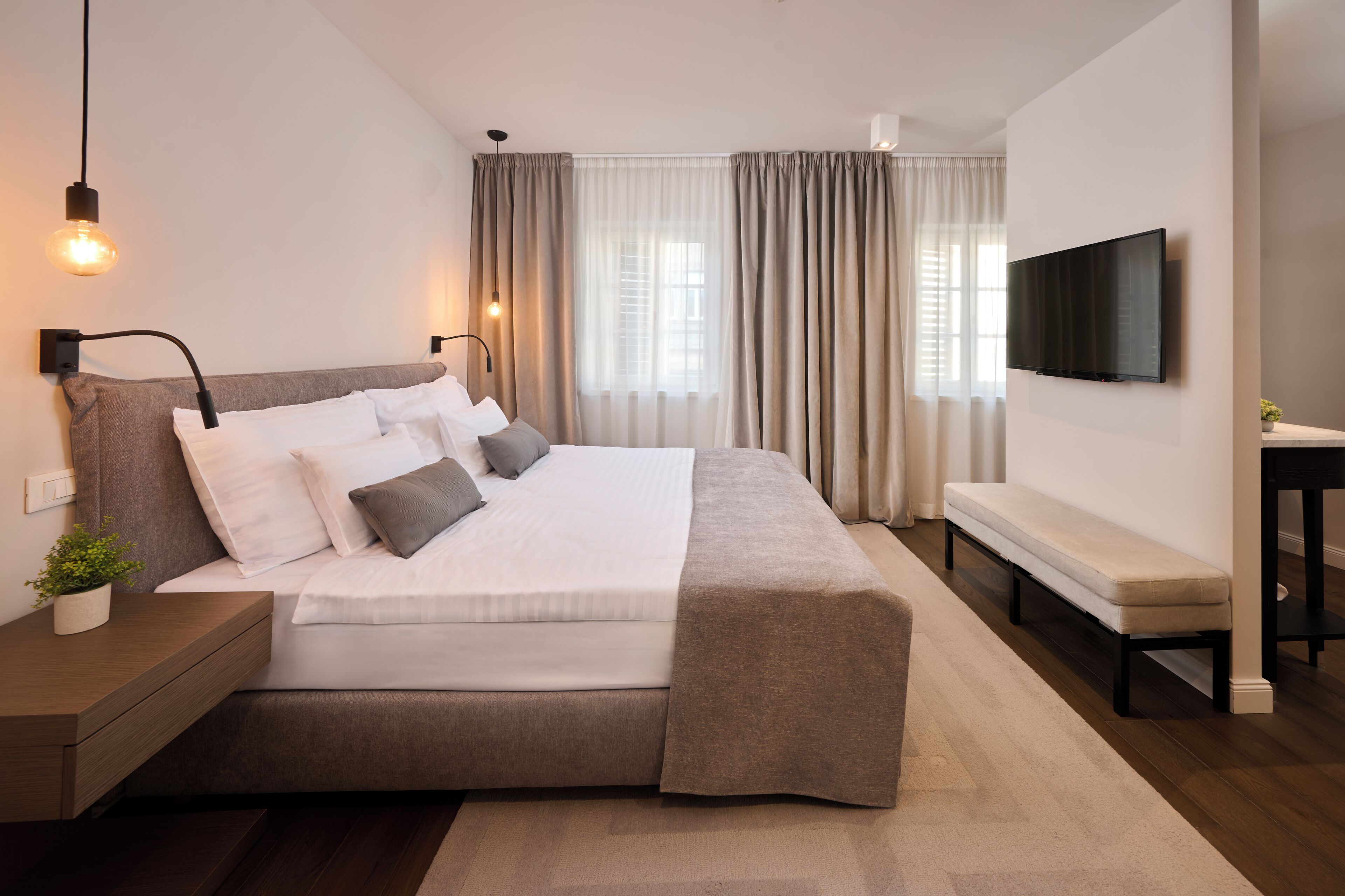 A modern bedroom with a bed, bedside table, bench, curtains, and a TV mounted on the wall.