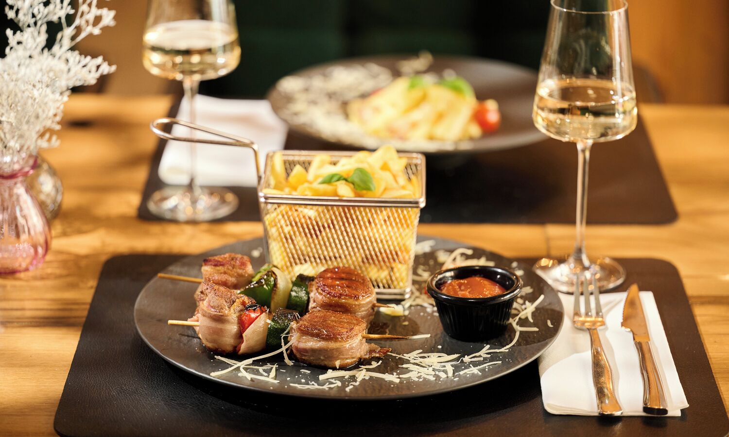 Una mesa de comedor con brochetas de carne y verduras, una copa de vino blanco y un recipiente con salsa.