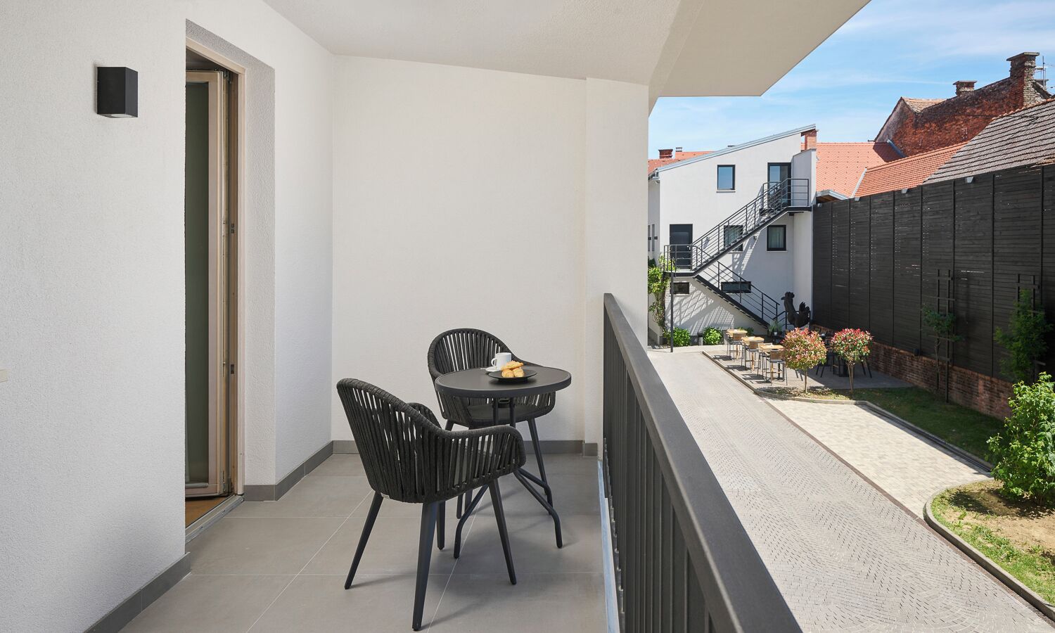 Un balcon avec une table et des chaises, une porte et une vue sur un immeuble avec un toit rouge.