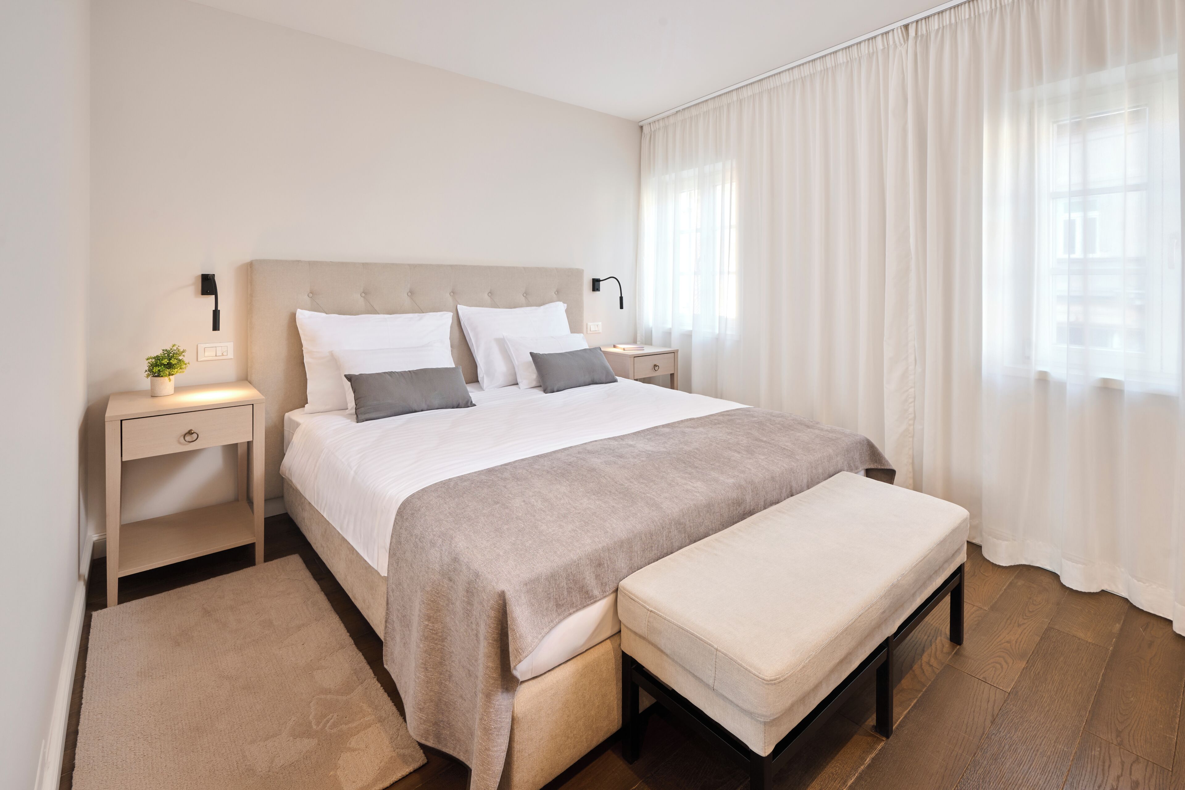 Un dormitorio ordenado con cama, mesitas de noche, un banco y una ventana con cortinas.