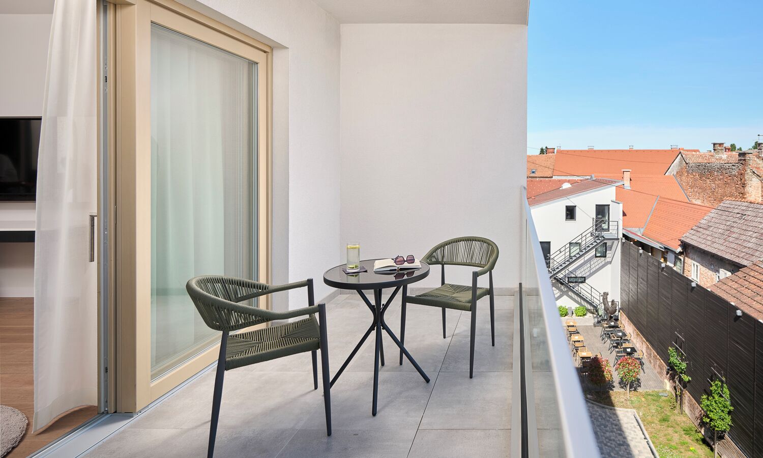 Un balcón con dos sillones de mimbre, una pequeña mesa, un ventanal corredero y vistas a un paisaje urbano con edificios y cielo despejado.