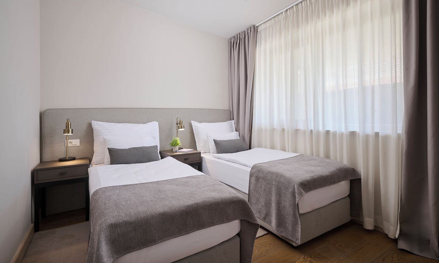 A hotel room with two beds, a nightstand with a lamp, a small table with a plant, and a window with curtains.