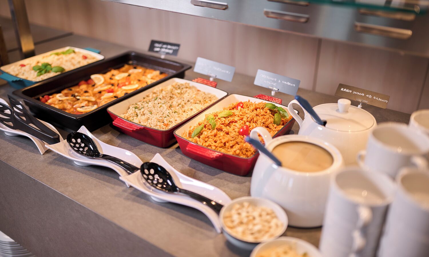 Un bufé con diversos platos, utensilios de servicio y tazas de café.