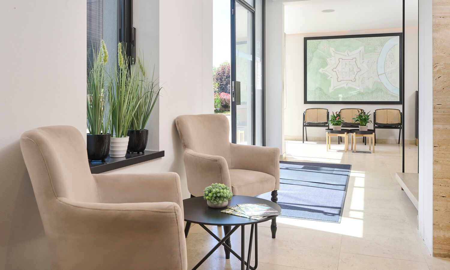 Een modern interieur met twee beige fauteuils, een tafeltje met een plant, en een eethoek met rieten stoelen en een kaart aan de muur.