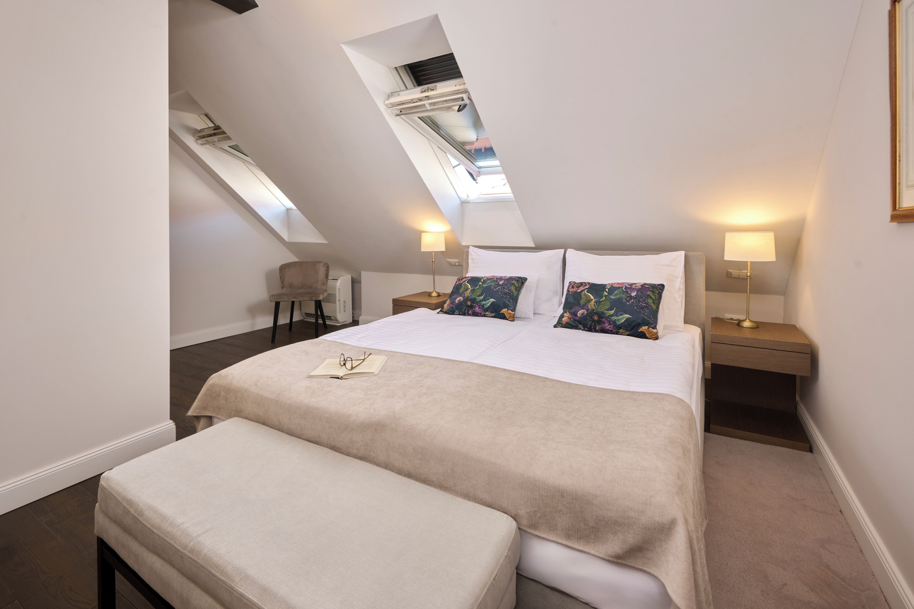 A bedroom with a skylight, a bed with a beige bedspread, two bedside tables with lamps, and a small chair.