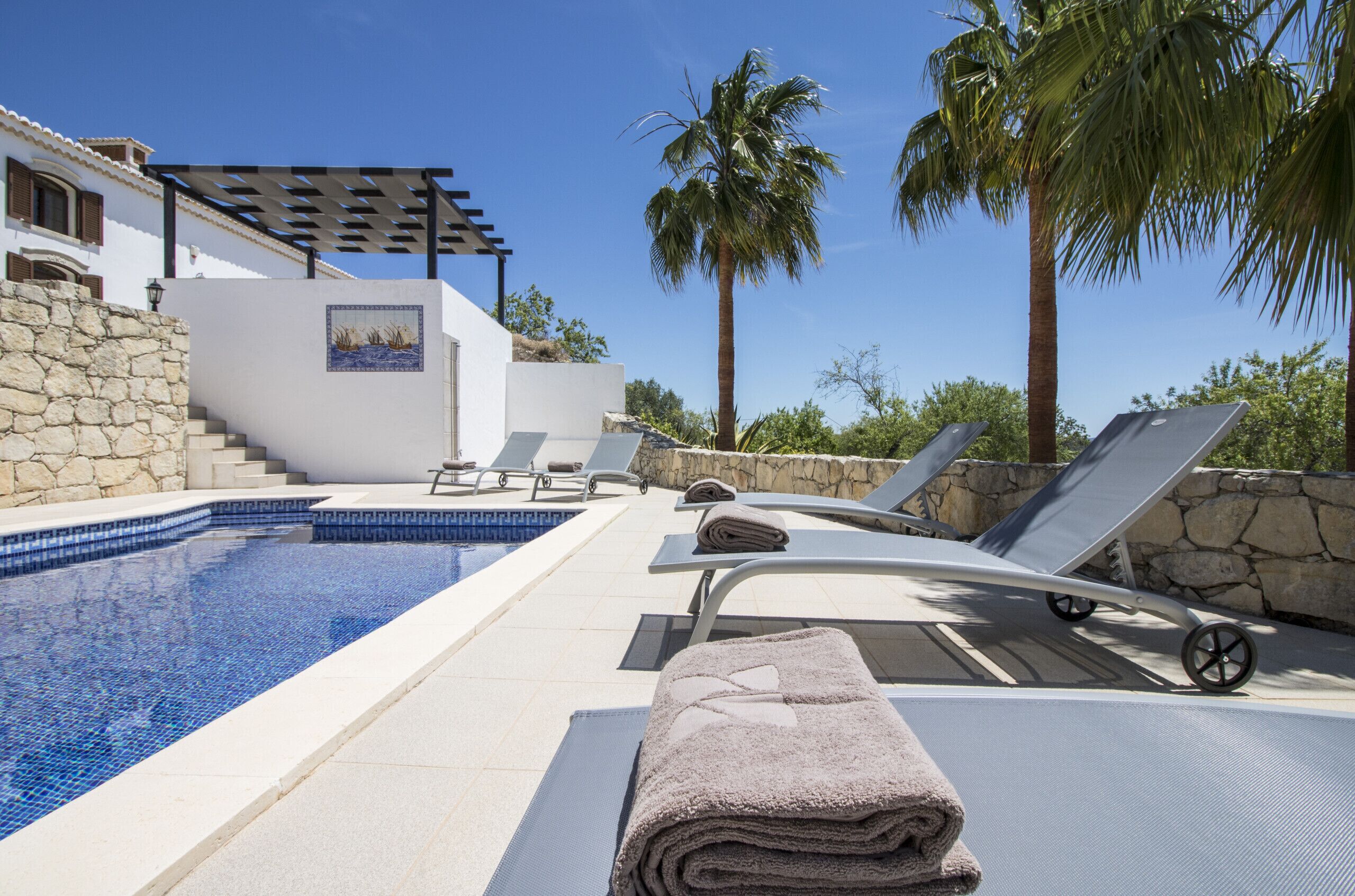 A pool area with lounge chairs, a pergola, and a stone wall.