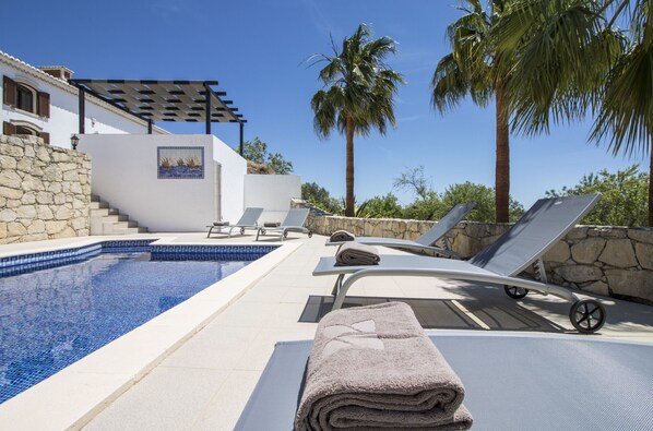A pool area with lounge chairs, a pergola, and a stone wall.