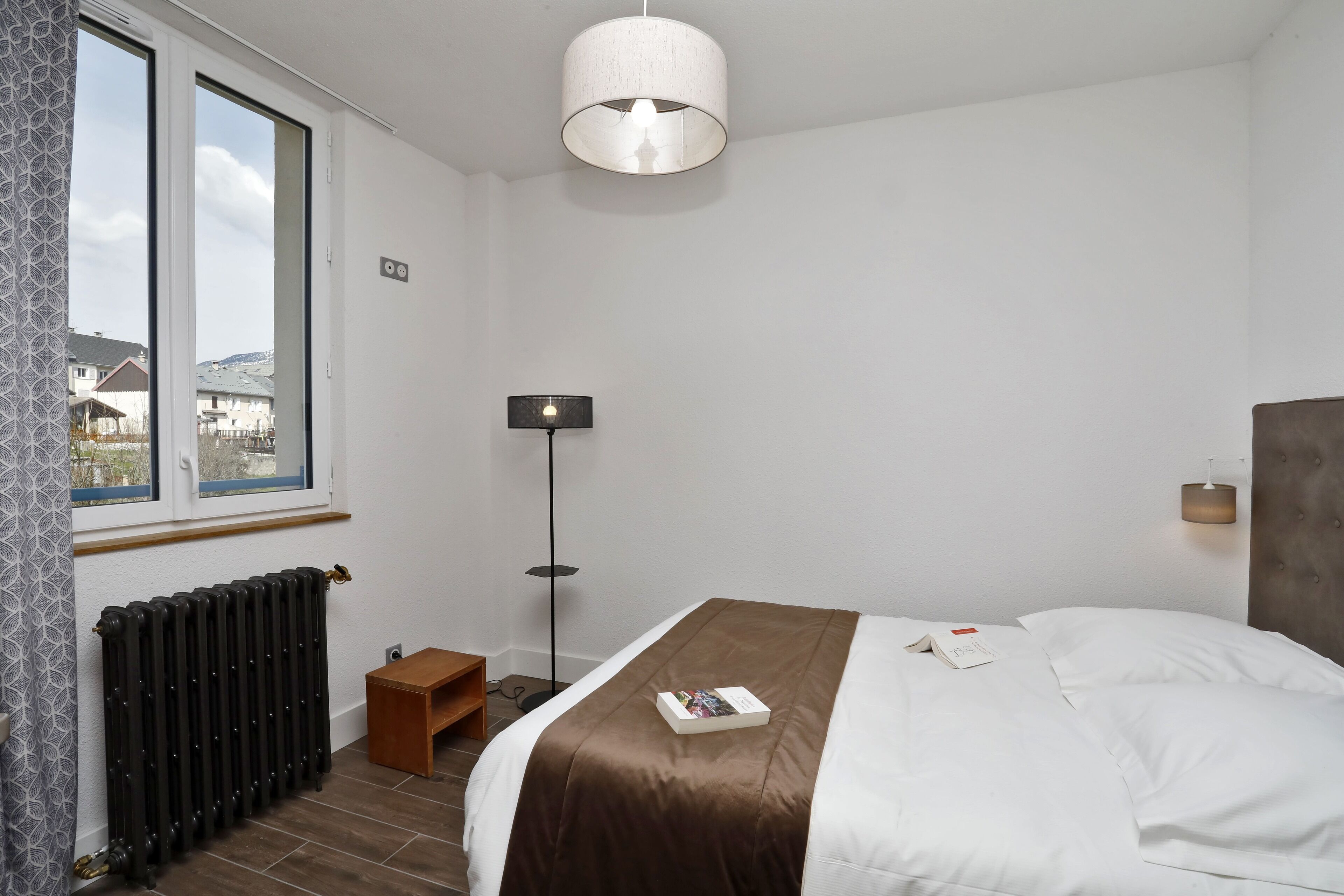 Une chambre à coucher avec un lit, un radiateur, une lampe sur pied et une fenêtre donnant sur des maisons.