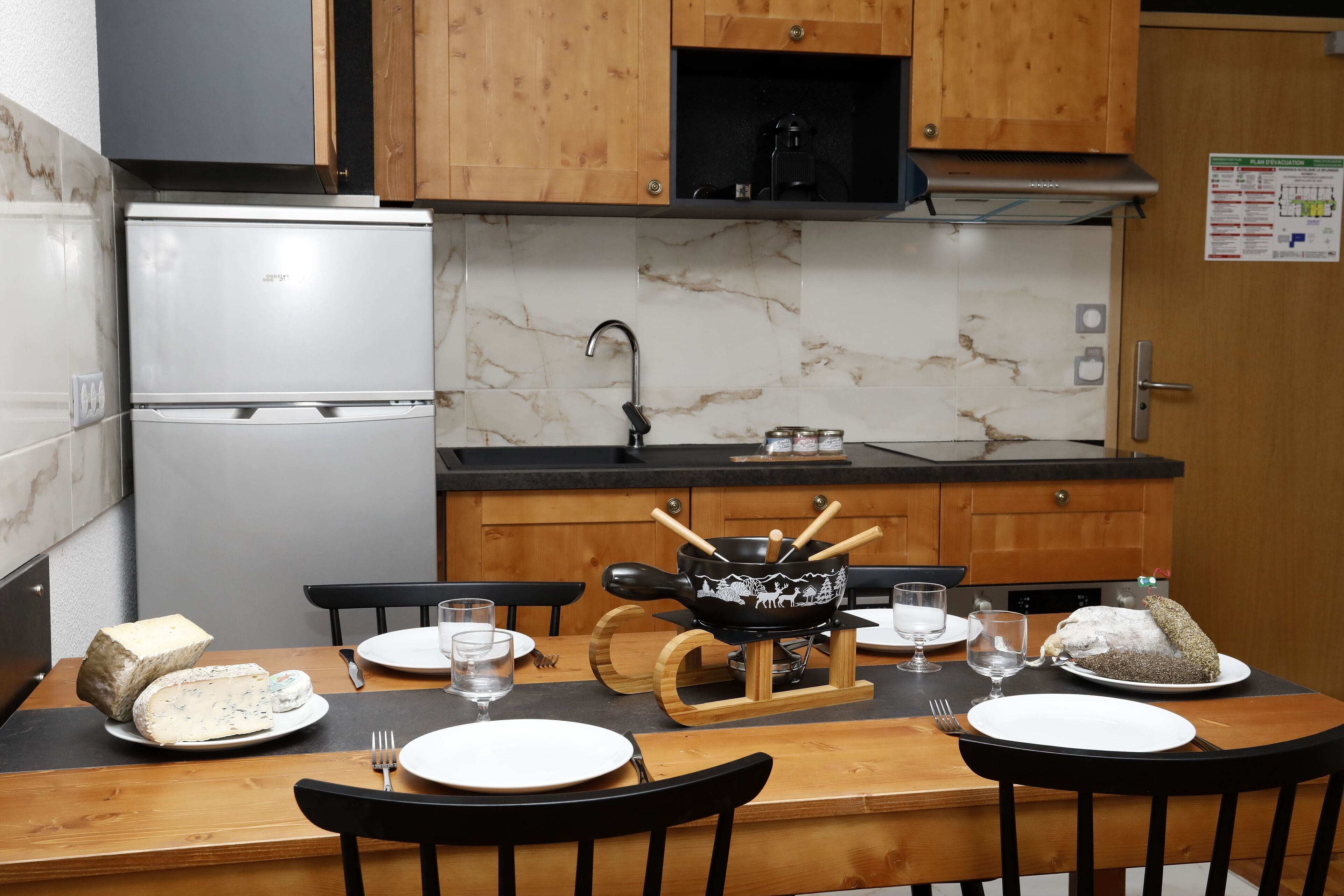 A kitchen with wooden cabinets, a white refrigerator, a marble backsplash, and a dining table set with plates, glasses, and a wooden serving board.