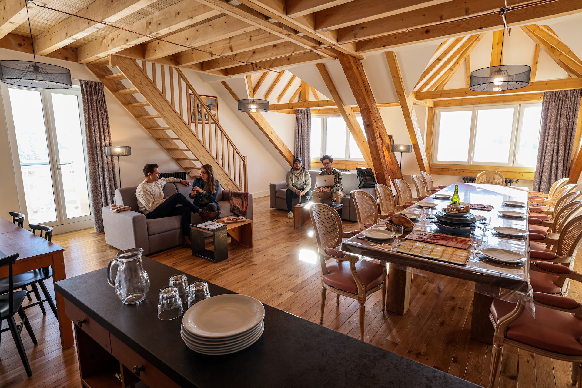 Un loft spacieux avec de hauts plafonds, des poutres apparentes, un coin repas avec une longue table et des chaises, et un coin salon confortable avec des canapés.