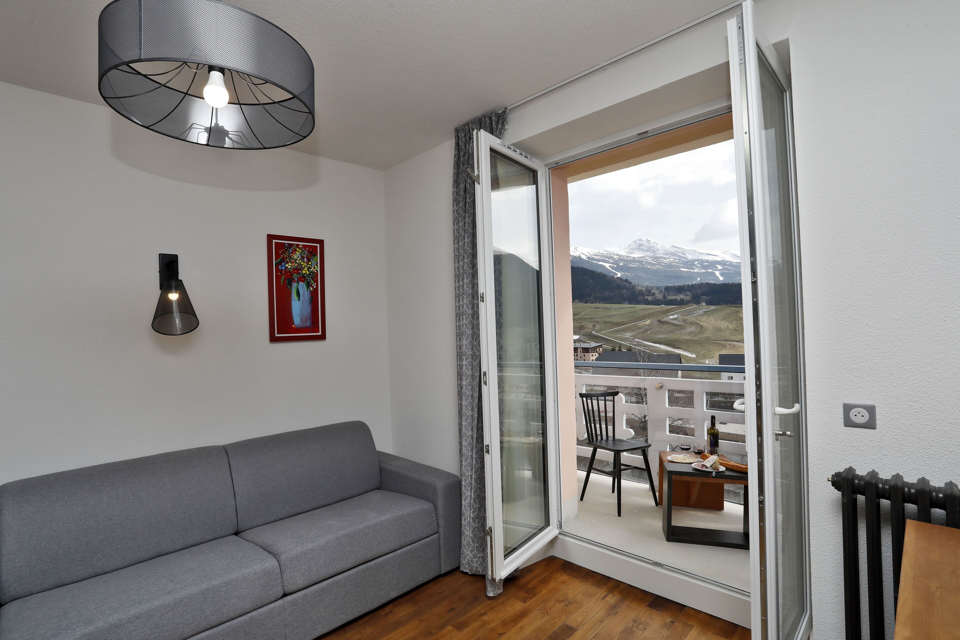 Ett modernt vardagsrum med en grå soffa, en hänglampa, ett inramat konstverk och en skjutdörr i glas som leder ut till en balkong med utsikt över snöklädda berg.