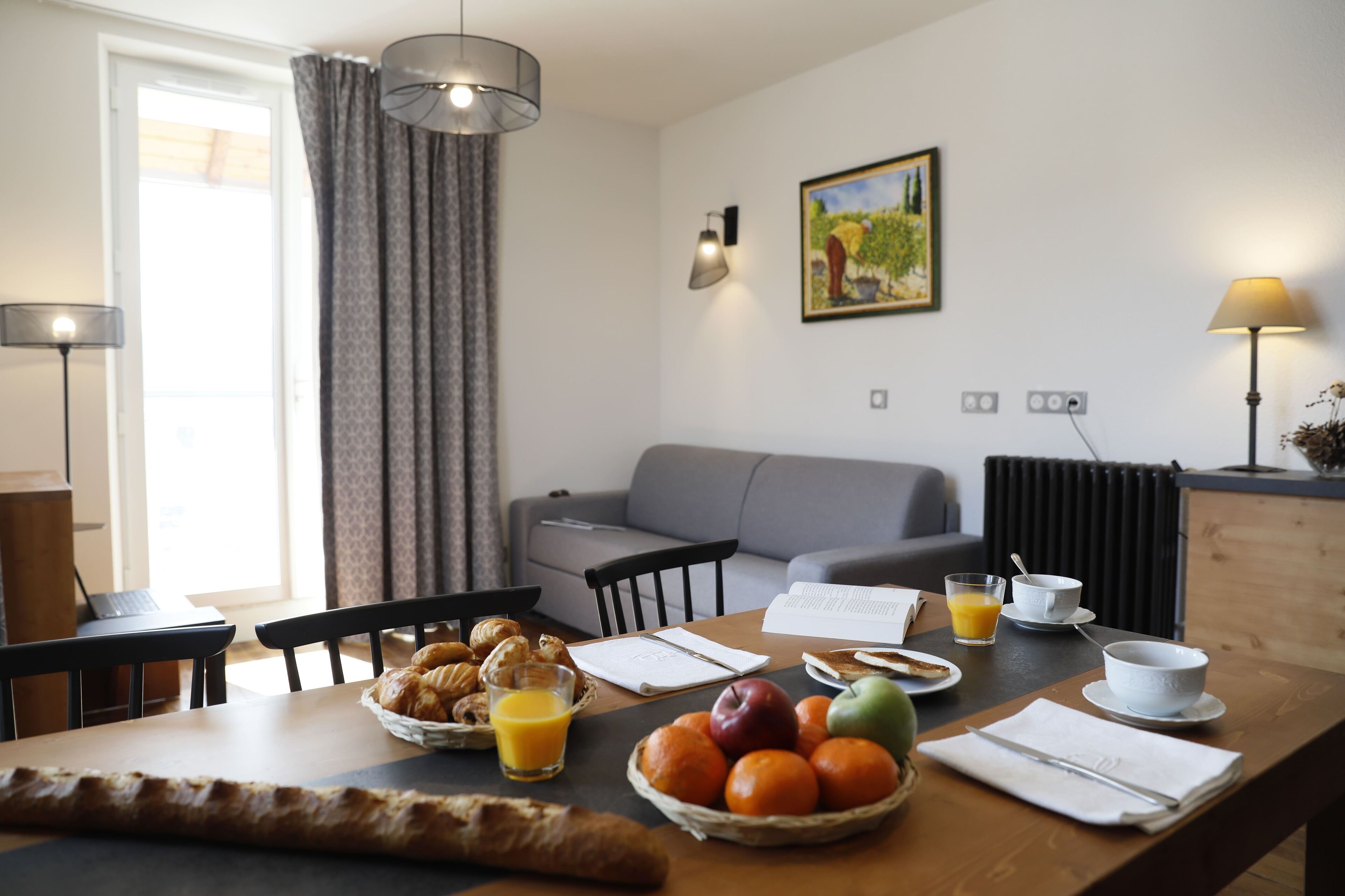 A breakfast table with croissants, fruit, and orange juice in a modern room with a sofa and a painting on the wall.
