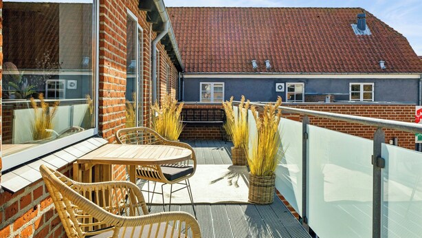 Een balkon met rieten meubilair, een glazen hekwerk en uitzicht op een gebouw met een rode dakbedekking.