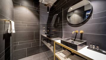 A modern bathroom with dark tiles, a round mirror, a glass-enclosed shower, and a gold-framed vanity.