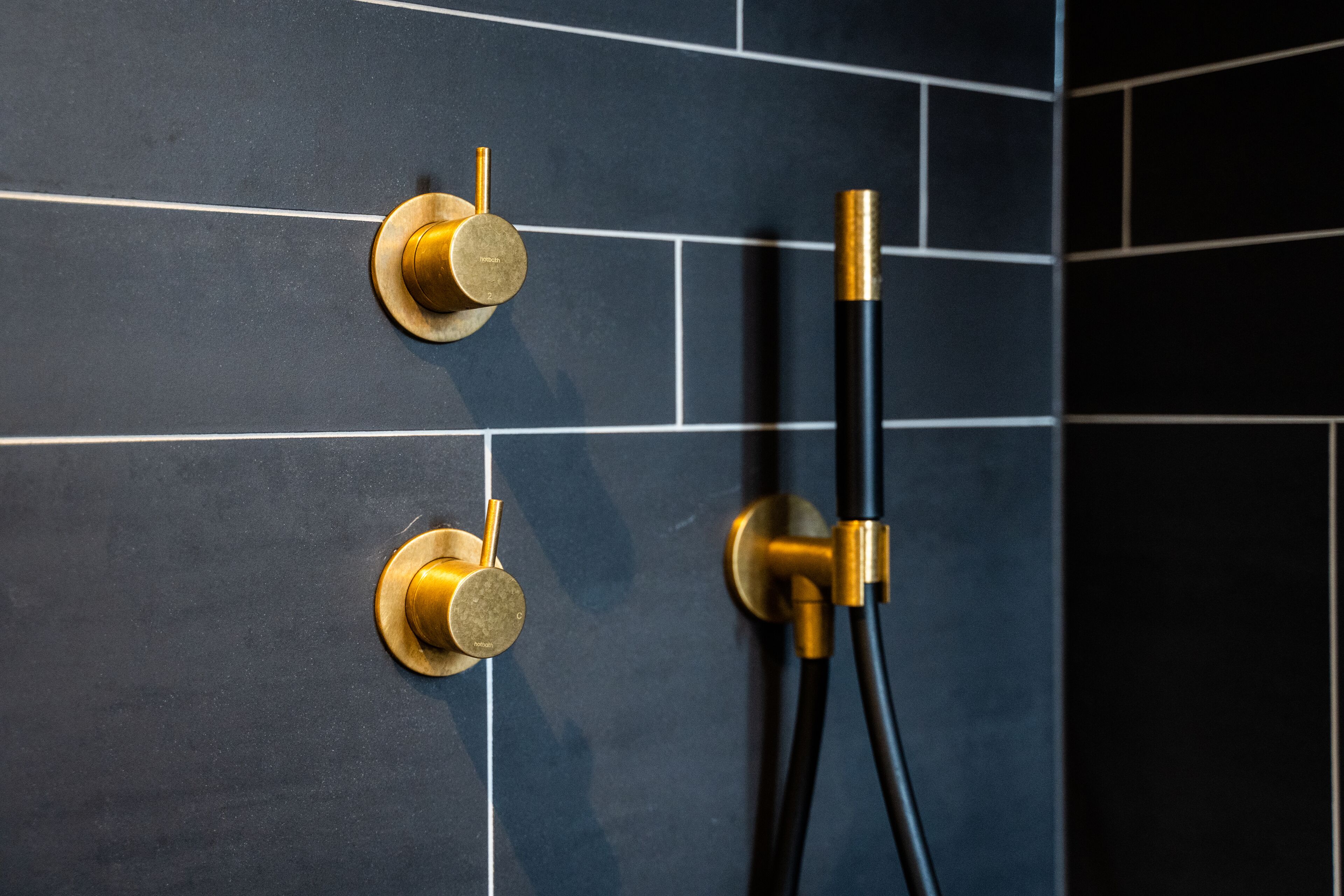 Ein Badezimmer mit dunkel gefliester Wand und einer goldfarbenen Duschsteuerung.
