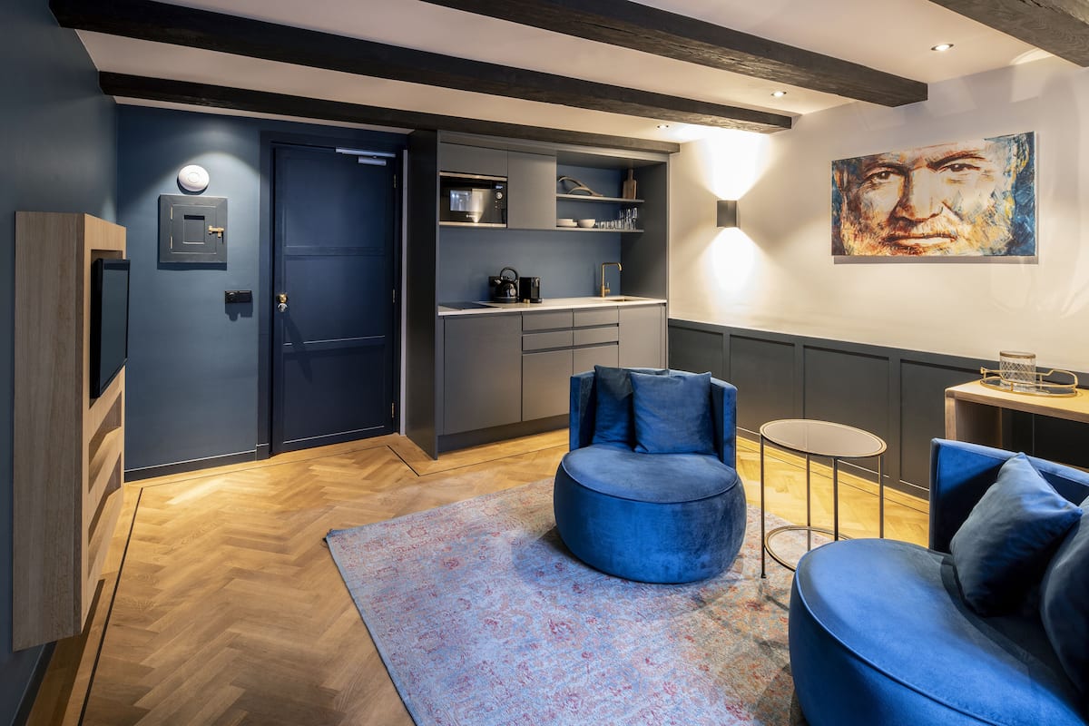 A modern living room with a blue sofa, a round blue chair, and a small round table. There is a kitchen area with a microwave and a coffee machine. The room has a wooden floor and a large painting on the wall.