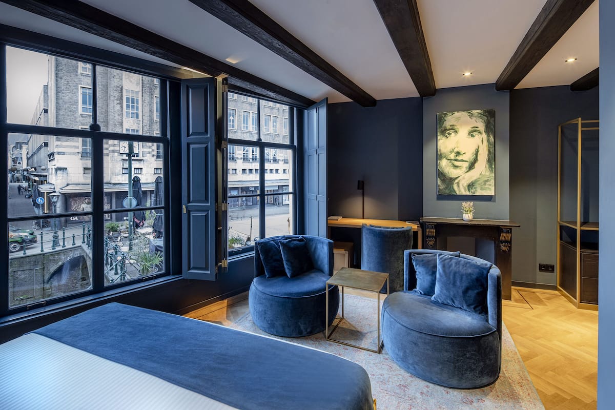 A modern room with large windows, a bed, two blue armchairs, and a small table.
