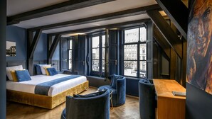 A modern bedroom with a large bed, a desk, and a chair. There are large windows and a view of buildings outside. - CTY Books (Leiden)