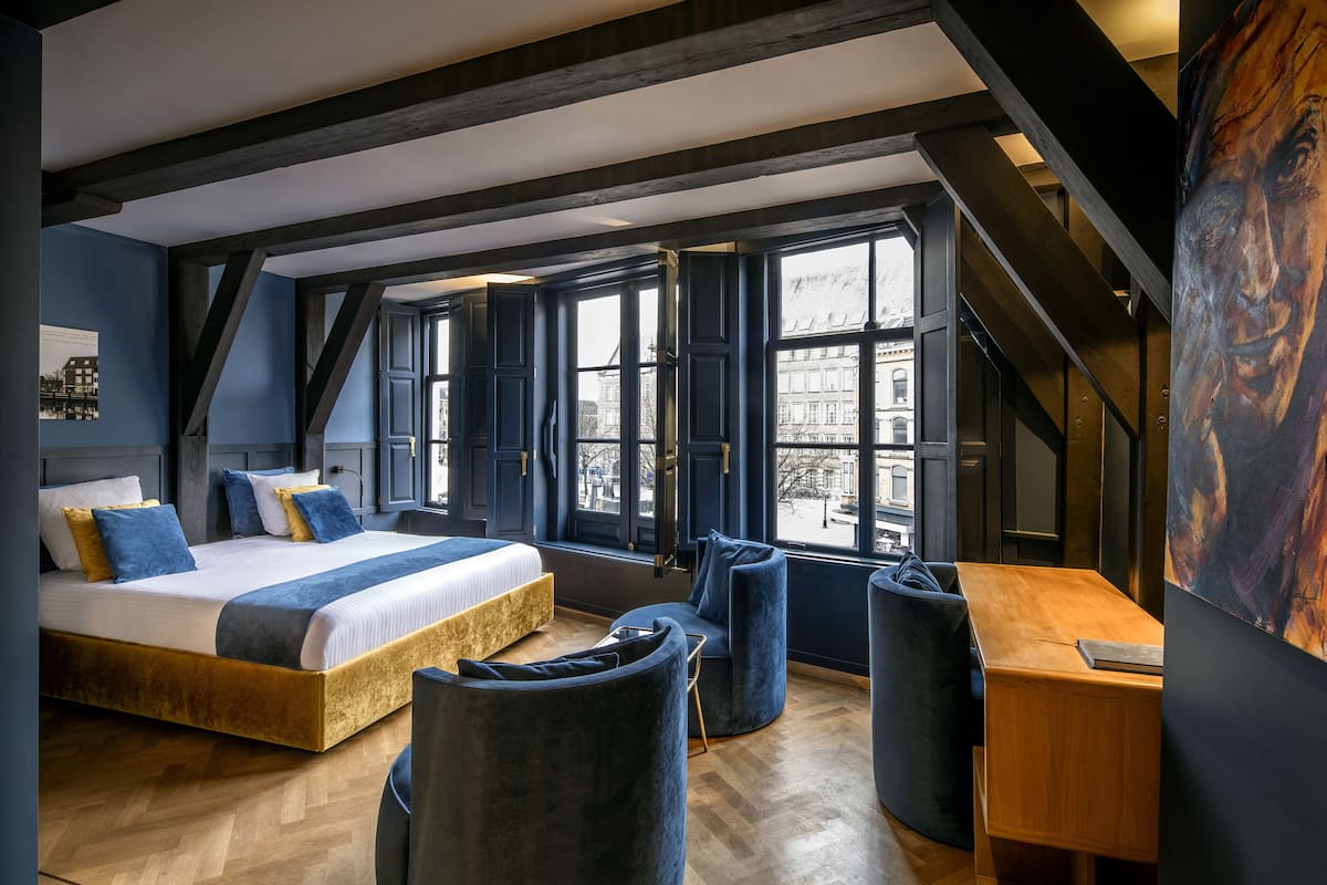 A modern bedroom with a large bed, a desk, and a chair. There are large windows and a view of buildings outside.