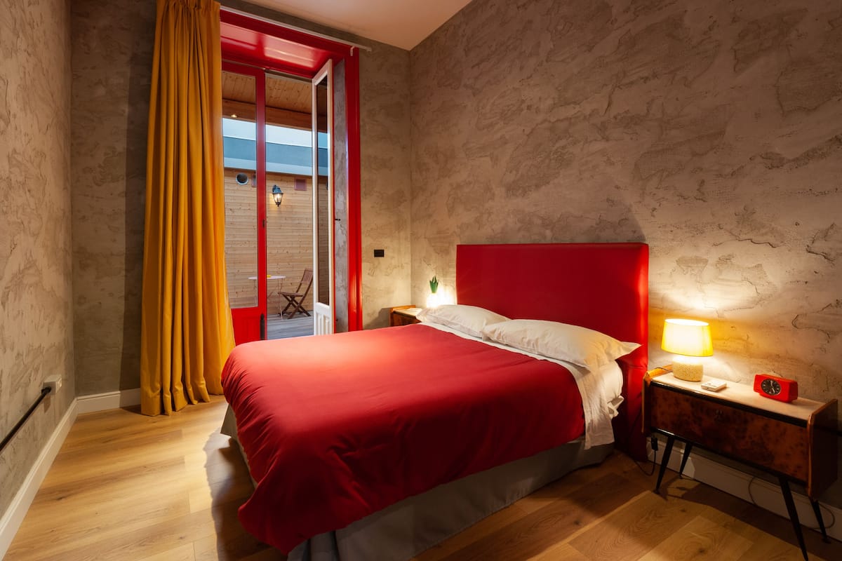 Une chambre avec un lit rouge, un plancher en bois, une tête de lit rouge, une table de chevet avec une lampe et un réveil rouge.