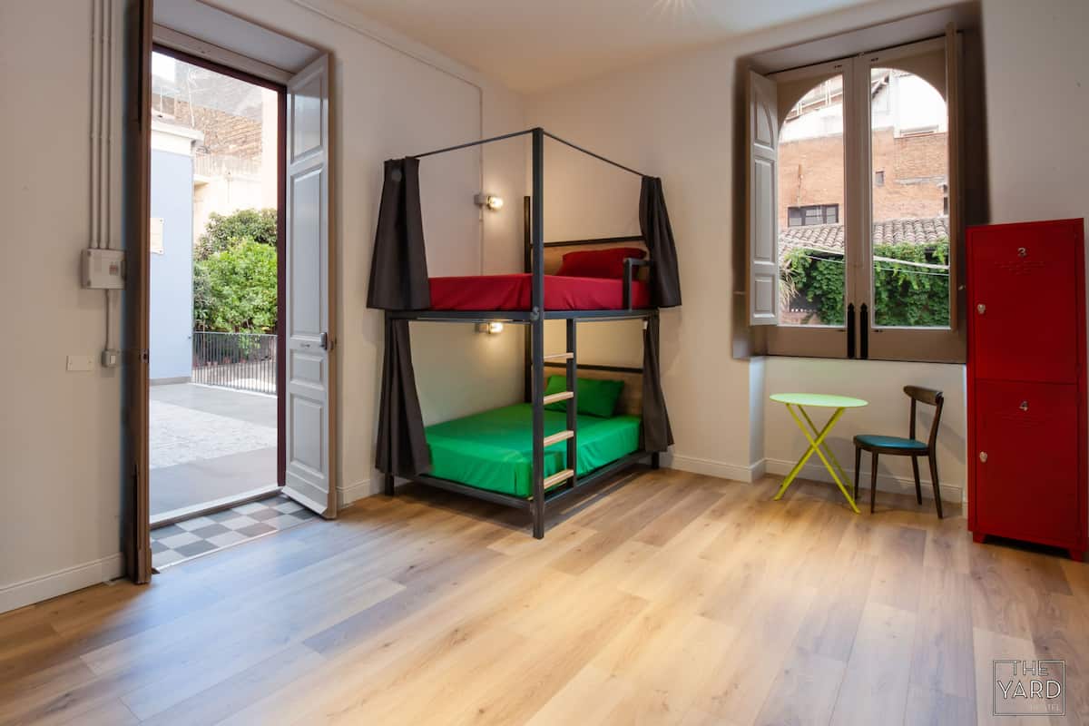 Une pièce avec un lit superposé, une petite table, une chaise, une armoire rouge et une fenêtre donnant sur un bâtiment et de la verdure.