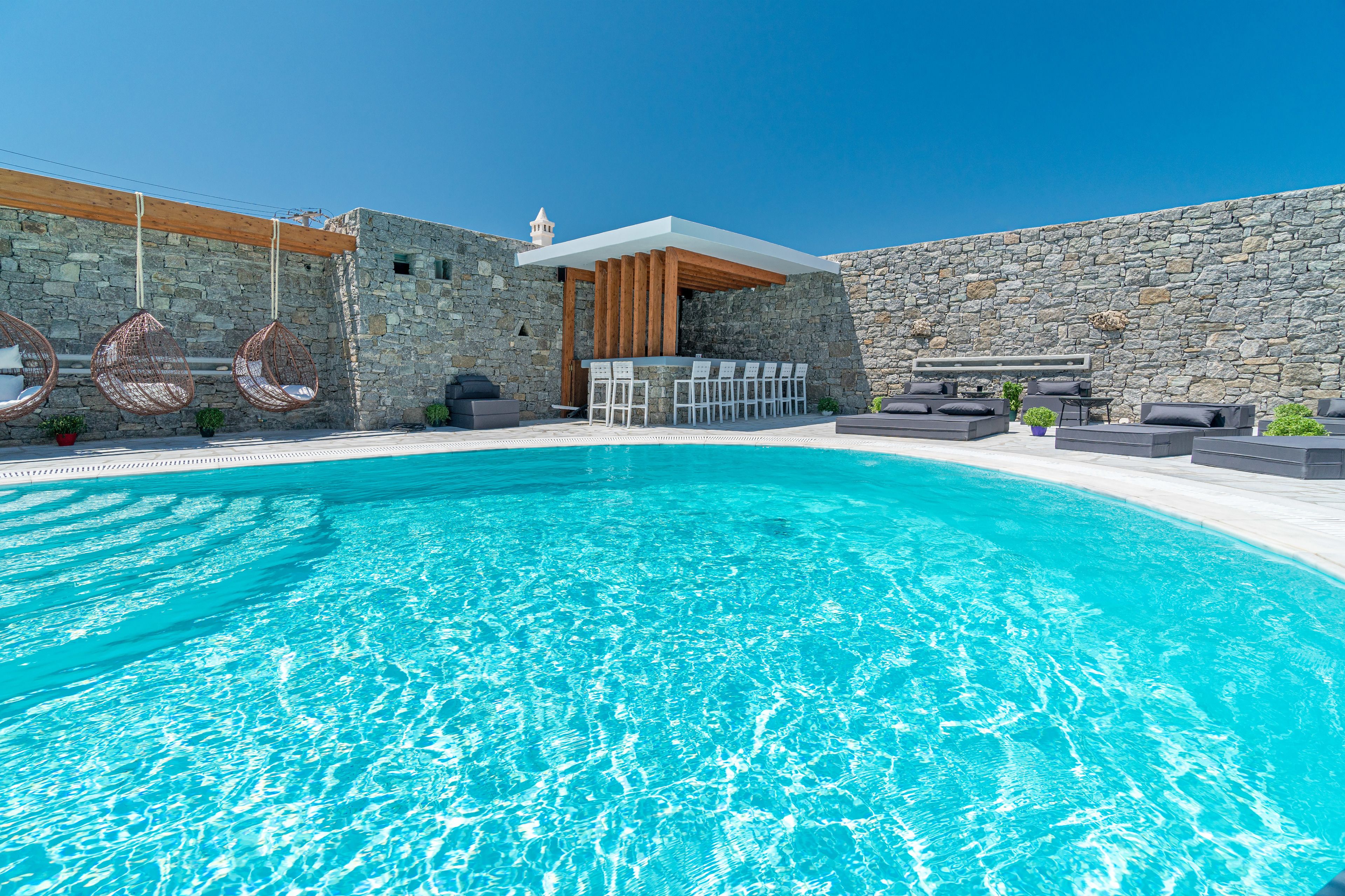 Zona junto a la piscina con muros de piedra, sillones colgantes y un área de estar cubierta con mesas y sillas.