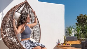 A person sitting in a wicker swing chair on a wooden deck with steps leading up to a white wall. - Omnia Mykonos Boutique Hotel & Suites (Mykonos)