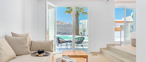 A modern living room with a sofa, a wooden table with a newspaper and a cup, and a view of an outdoor pool area through glass doors.