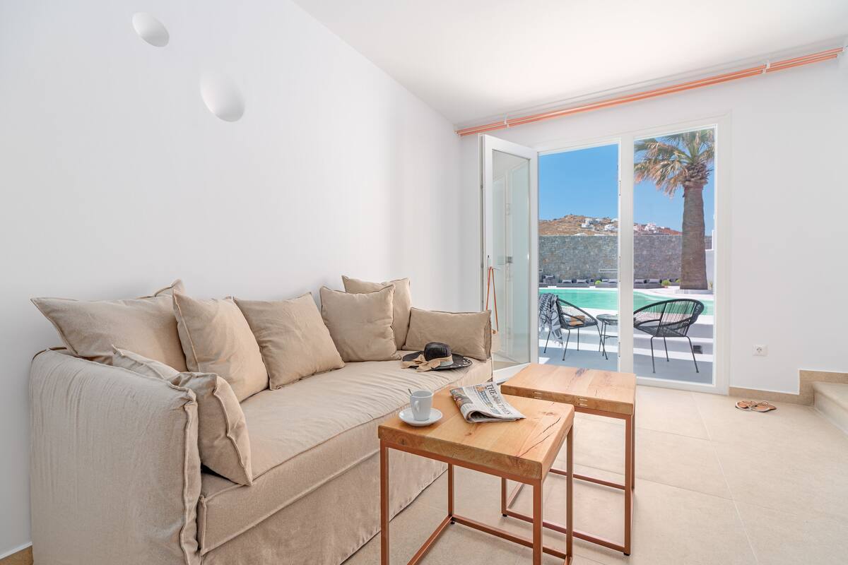 Ein modernes Wohnzimmer mit einem beigefarbenen Sofa, einem Holztisch und Blick auf einen Pool und eine Palme durch eine Schiebetür.