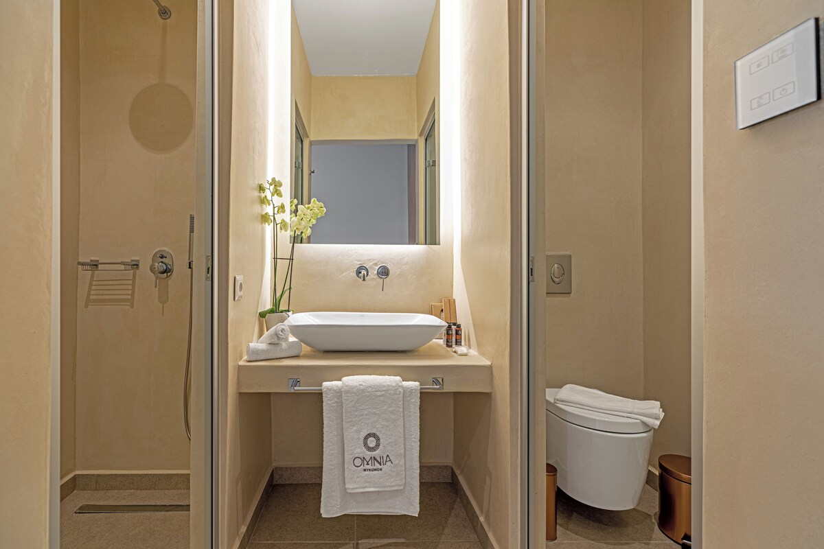 A bathroom with a white sink, a mirror, and a toilet. There is a towel hanging on the wall and a small plant on the sink.