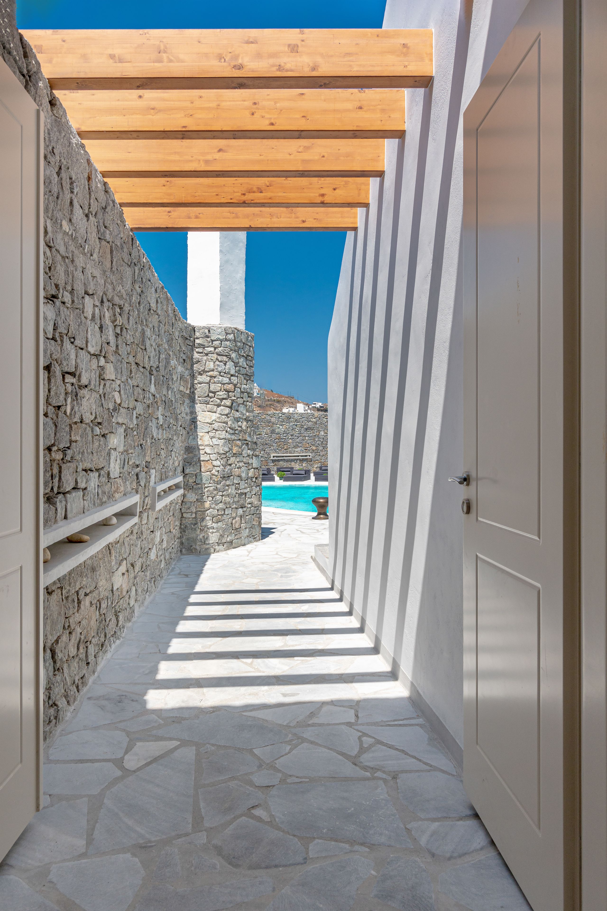 A narrow stone corridor with a wooden pergola overhead, leading to a pool area with a clear blue sky in the background.