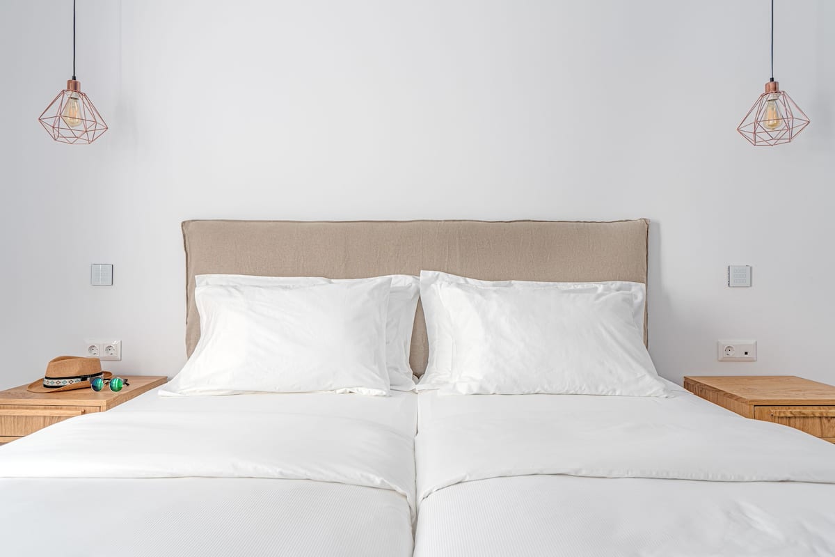 Ein ordentlich bezogenes Bett mit einem beige gefärbten Kopfteil, weißen Kissen und einem Nachttisch aus Holz, auf dem ein Hut und eine Sonnenbrille liegen.
