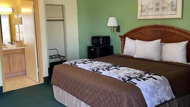 A hotel room with a bed, a nightstand, a lamp, a microwave, and a door leading to a bathroom.