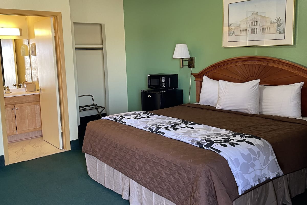 Habitación de hotel con cama, mesita de noche, lámpara, microondas y puerta que da a un baño.