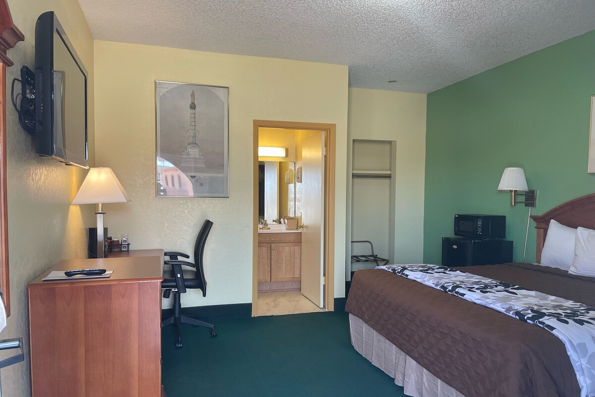 Habitación de hotel con cama, escritorio, silla, televisor y baño visible a través de una puerta abierta.