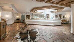 A spacious room with a stone floor, a central seating area with a cowhide rug, wooden beams on the ceiling, and a reception desk with a mirror and heart-shaped decorations. - Sporthotel Beck (Brand)