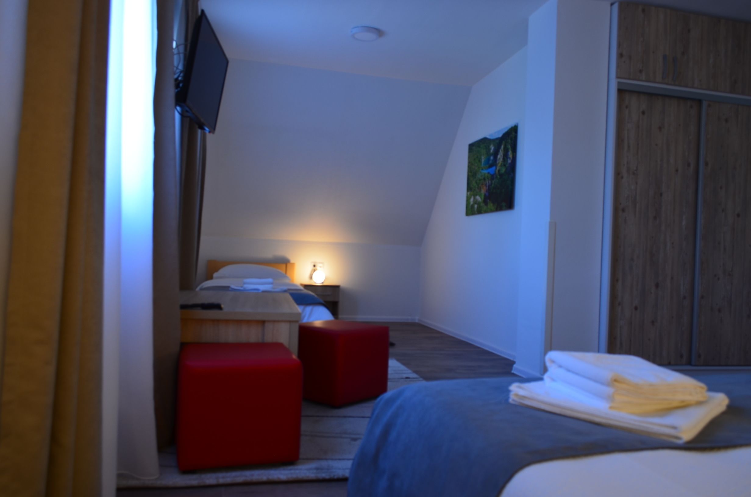 A modern bedroom with a bed, a red ottoman, a nightstand, a TV, and a wooden wardrobe.