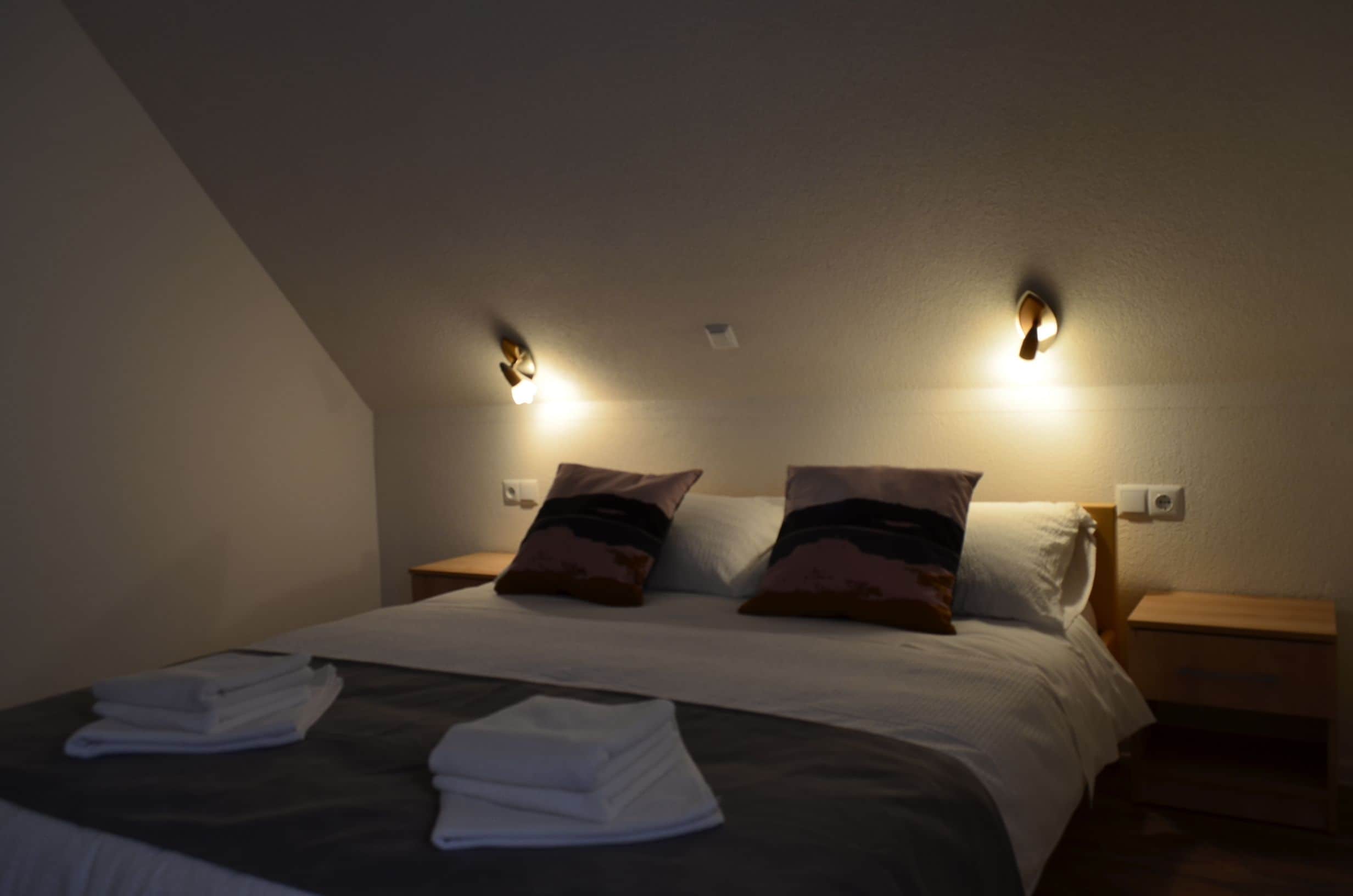 Een net opgemaakt bed met kussens, een nachtkastje en twee wandlampen.