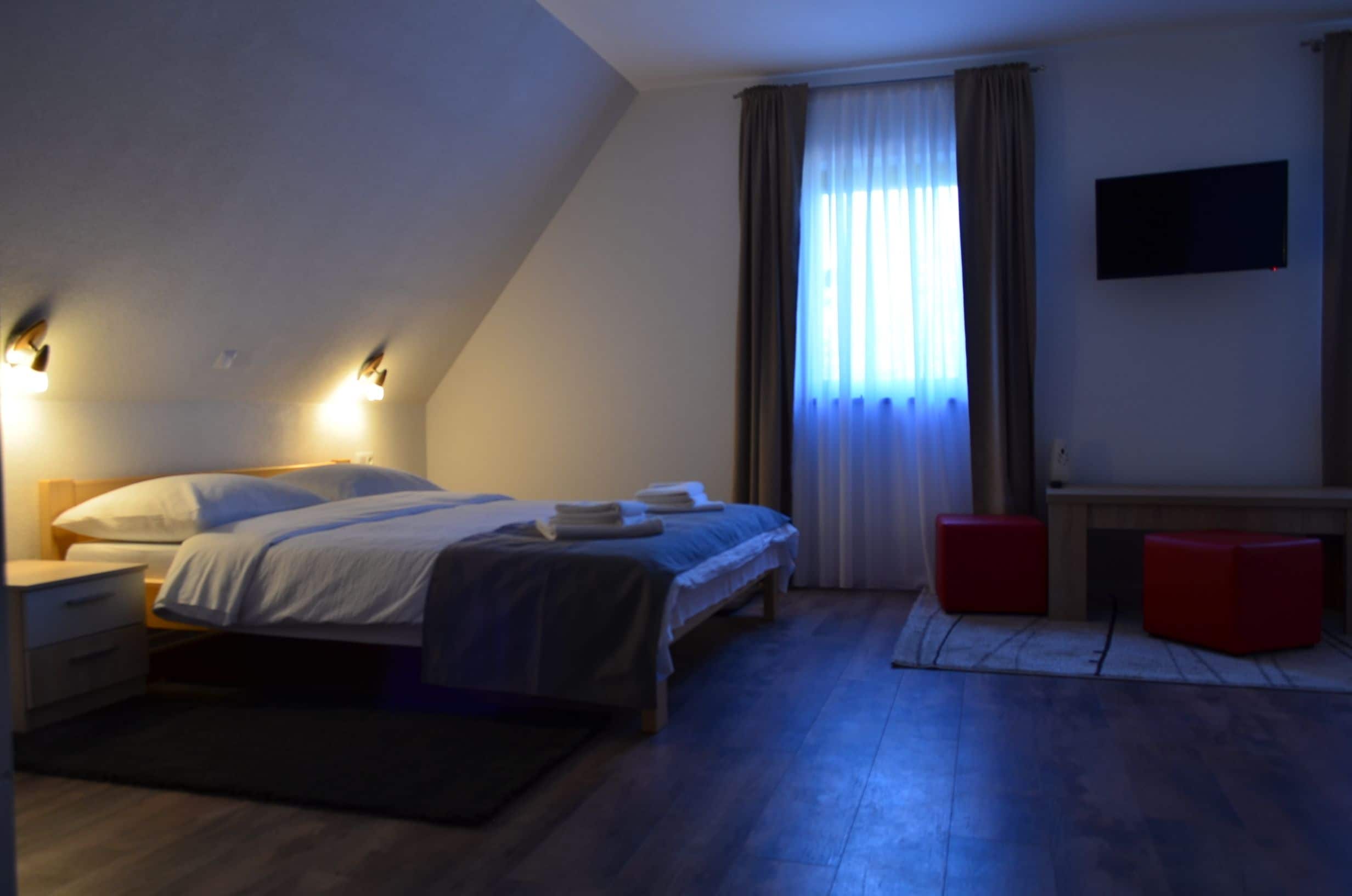 A bedroom with a bed, bedside tables, a flat-screen TV, and a window with curtains.