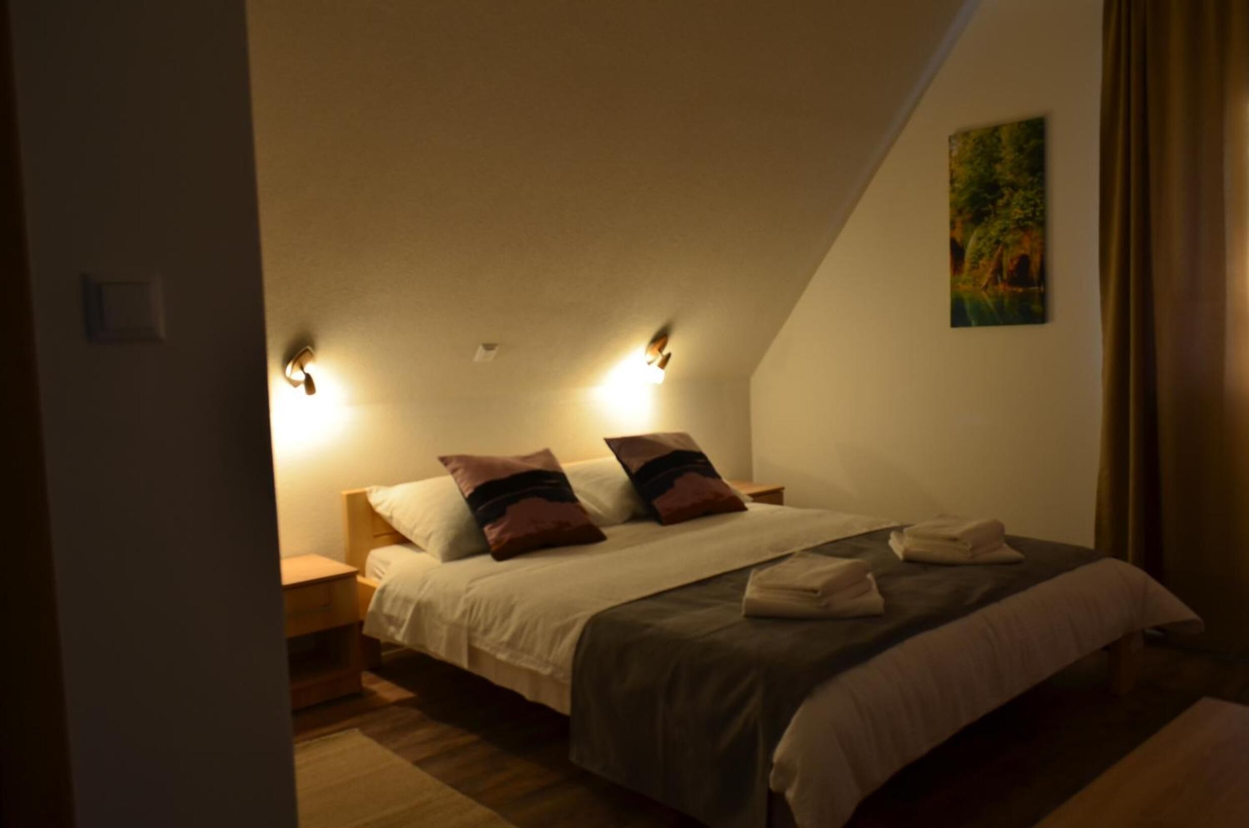 A bedroom with a sloped ceiling, two bedside lamps, a bed with pillows, and a painting on the wall.