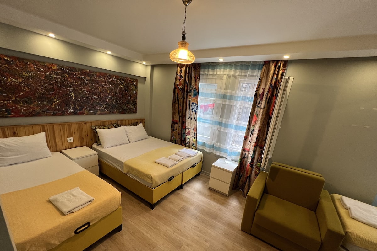 A hotel room with a large bed, a smaller bed, a sofa, a chair, a desk, and a large abstract painting on the wall.