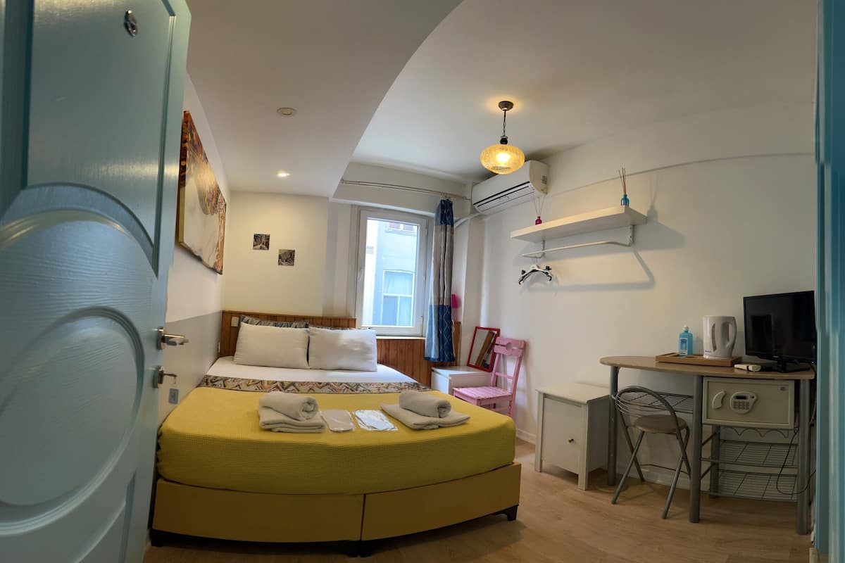 A compact hotel room with a bed, a desk with a computer, a chair, a small table, and a window.