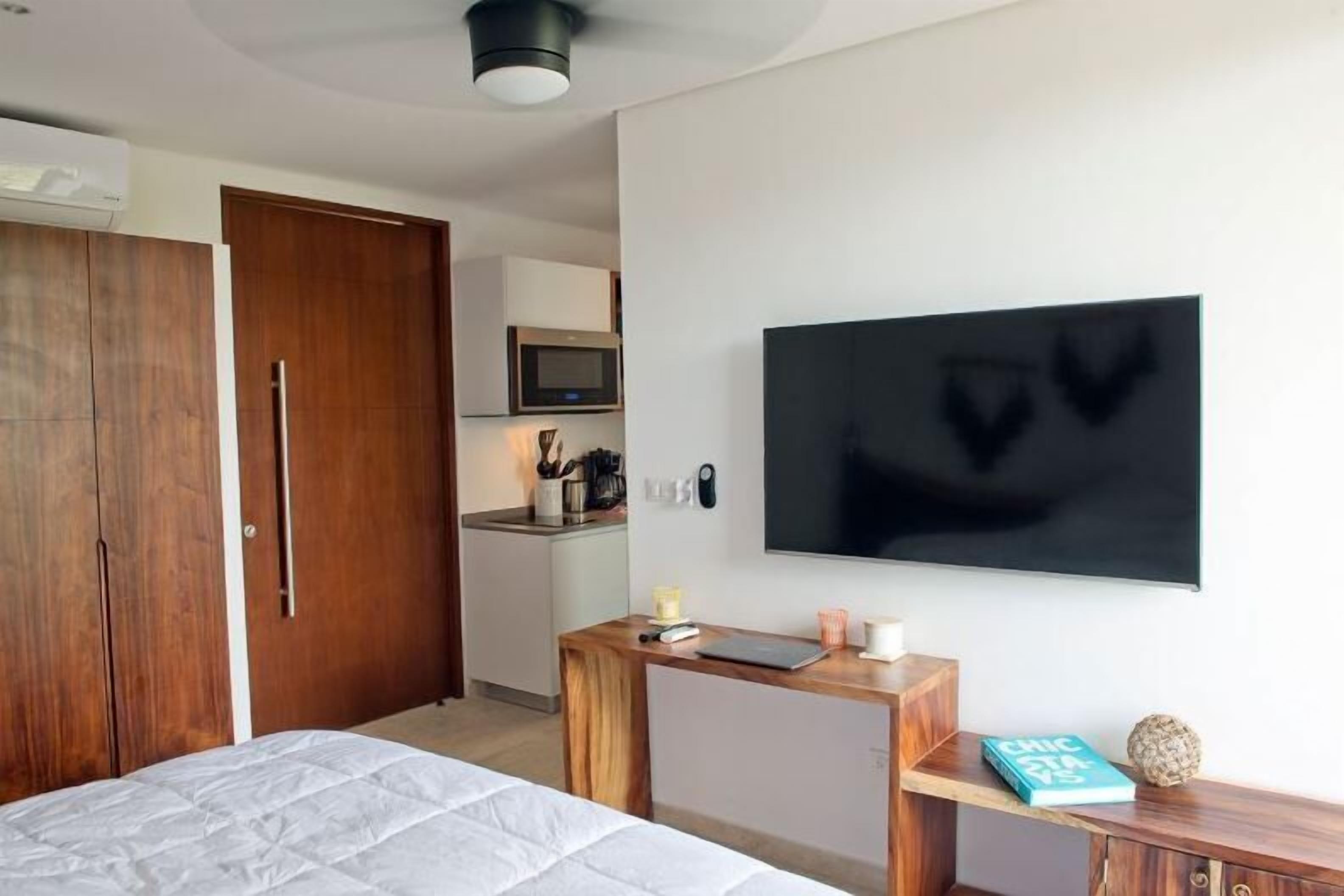 A modern hotel room with a bed, a wooden desk, a flat-screen TV, a wardrobe, and a kitchenette in the background.