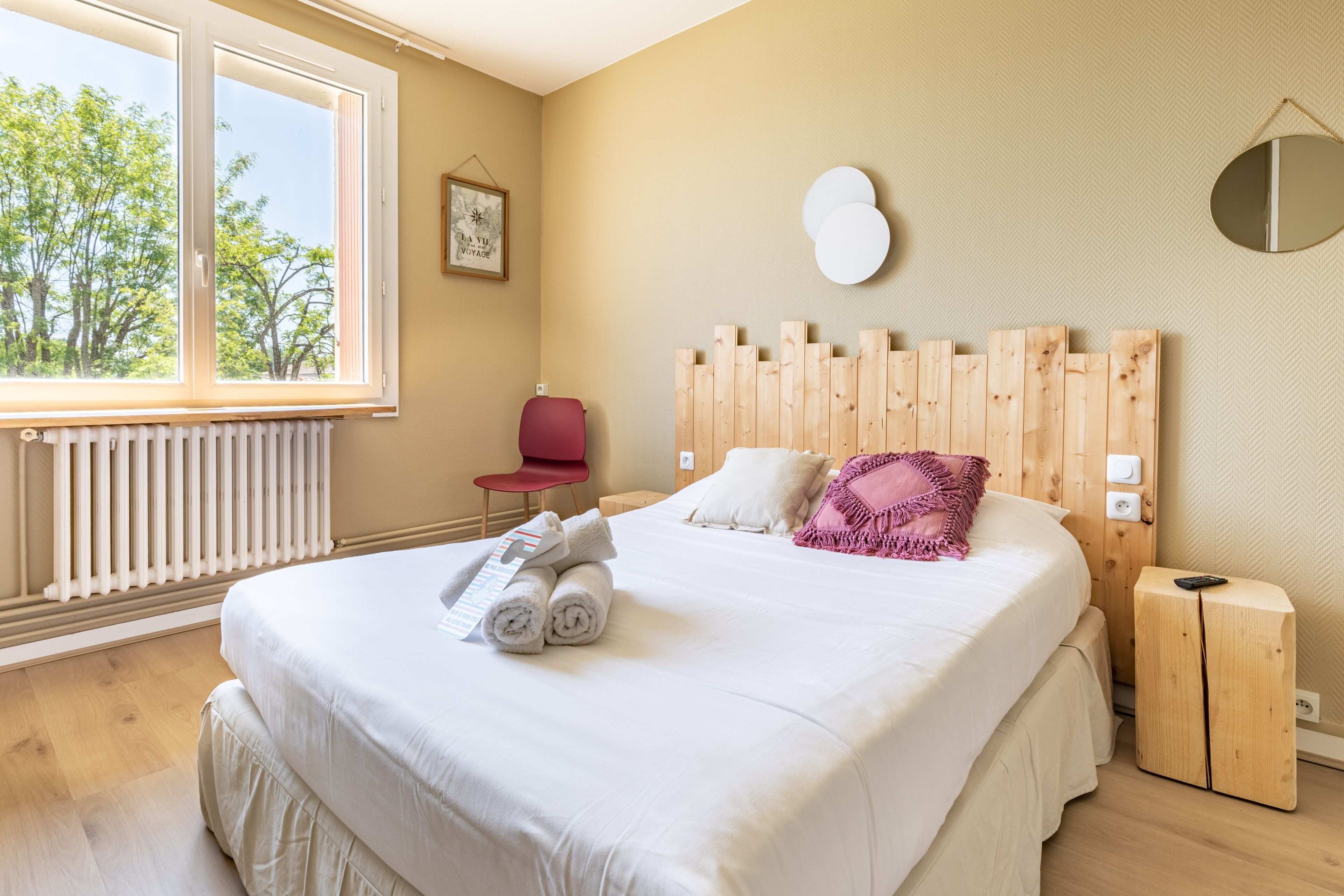 Ein Schlafzimmer mit großem Fenster, einem Holzkopfteil, einem Bett mit weißer Bettwäsche, einem roten Sessel und einem kleinen Nachttisch.