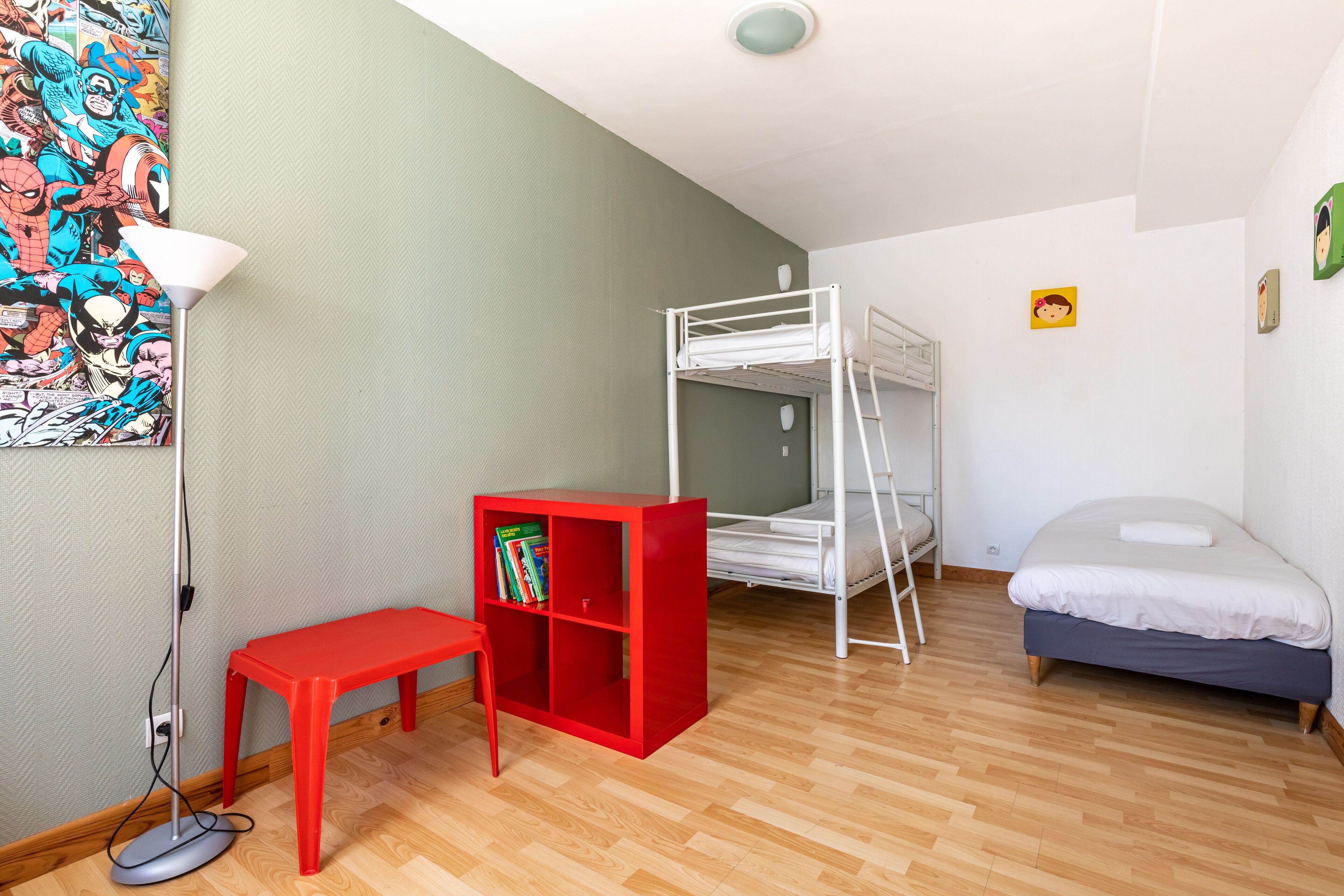 Een kamer met een stapelbed, een rode boekenkast, een klein tafeltje en een vloerlamp.