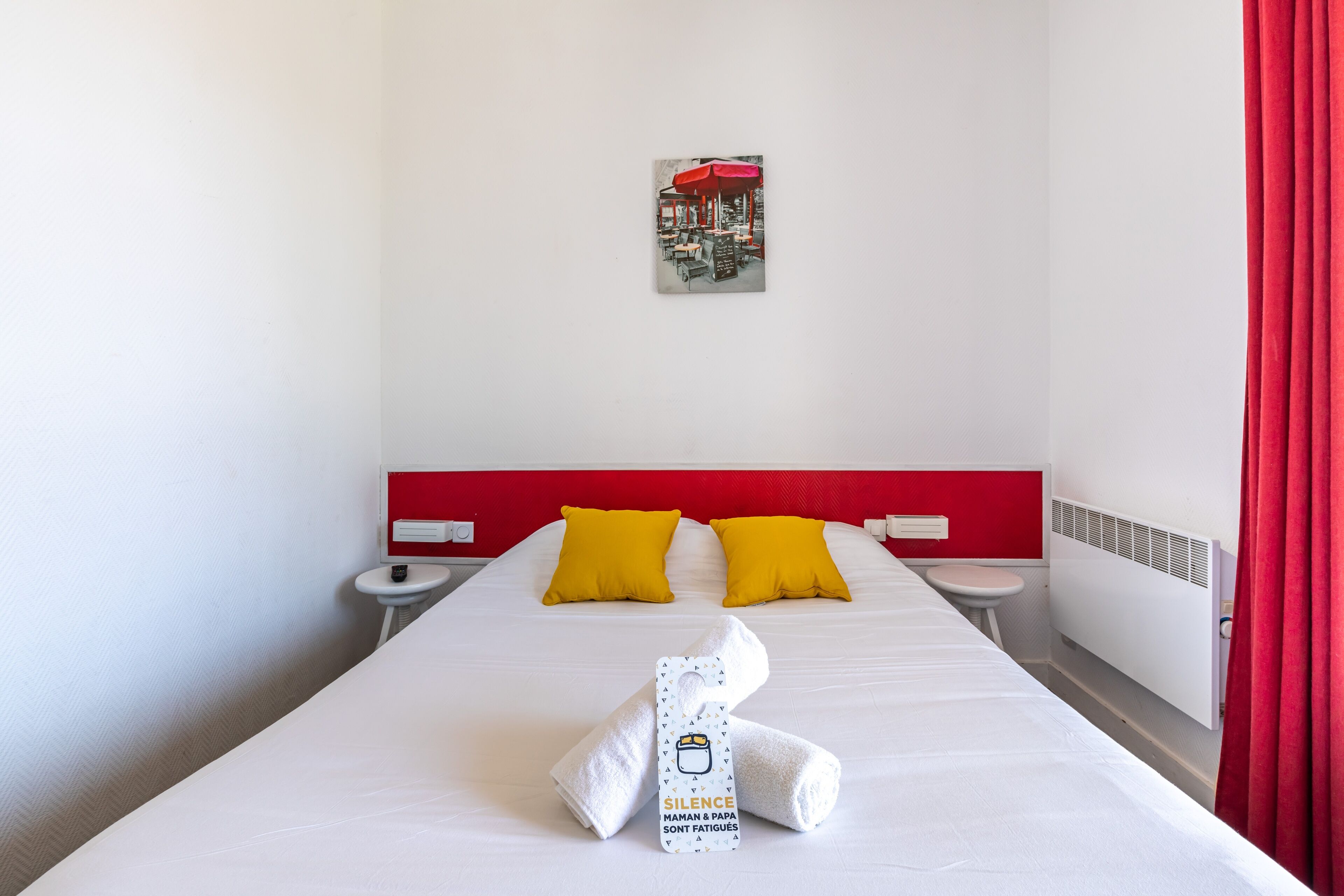 Habitación de hotel con cama blanca, cabecera roja, dos almohadas amarillas, mesita de noche blanca, cortina roja y una toalla con etiqueta.