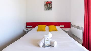 Habitación de hotel con cama blanca, cabecera roja, dos almohadas amarillas, mesita de noche blanca, cortina roja y una toalla con etiqueta.