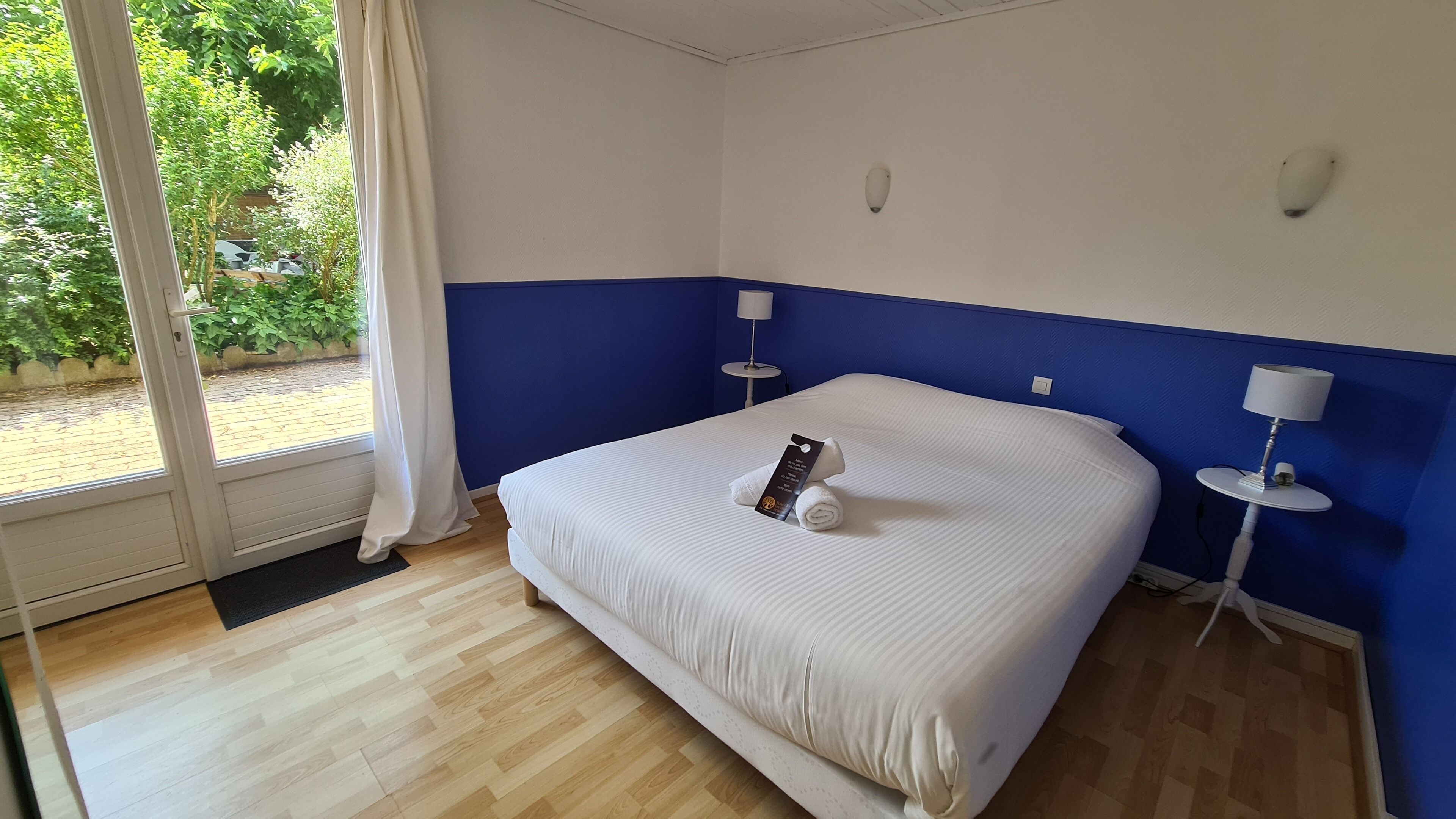A hotel room with a bed, a window with curtains, a bedside table with a lamp, and a view of greenery outside.