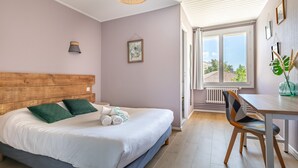 A bedroom with a wooden headboard, a bed with white bedding and green pillows, a bedside table with a lamp, and a view of the outdoors through a window. - Hotel les vieux Acacias (Queyrac)