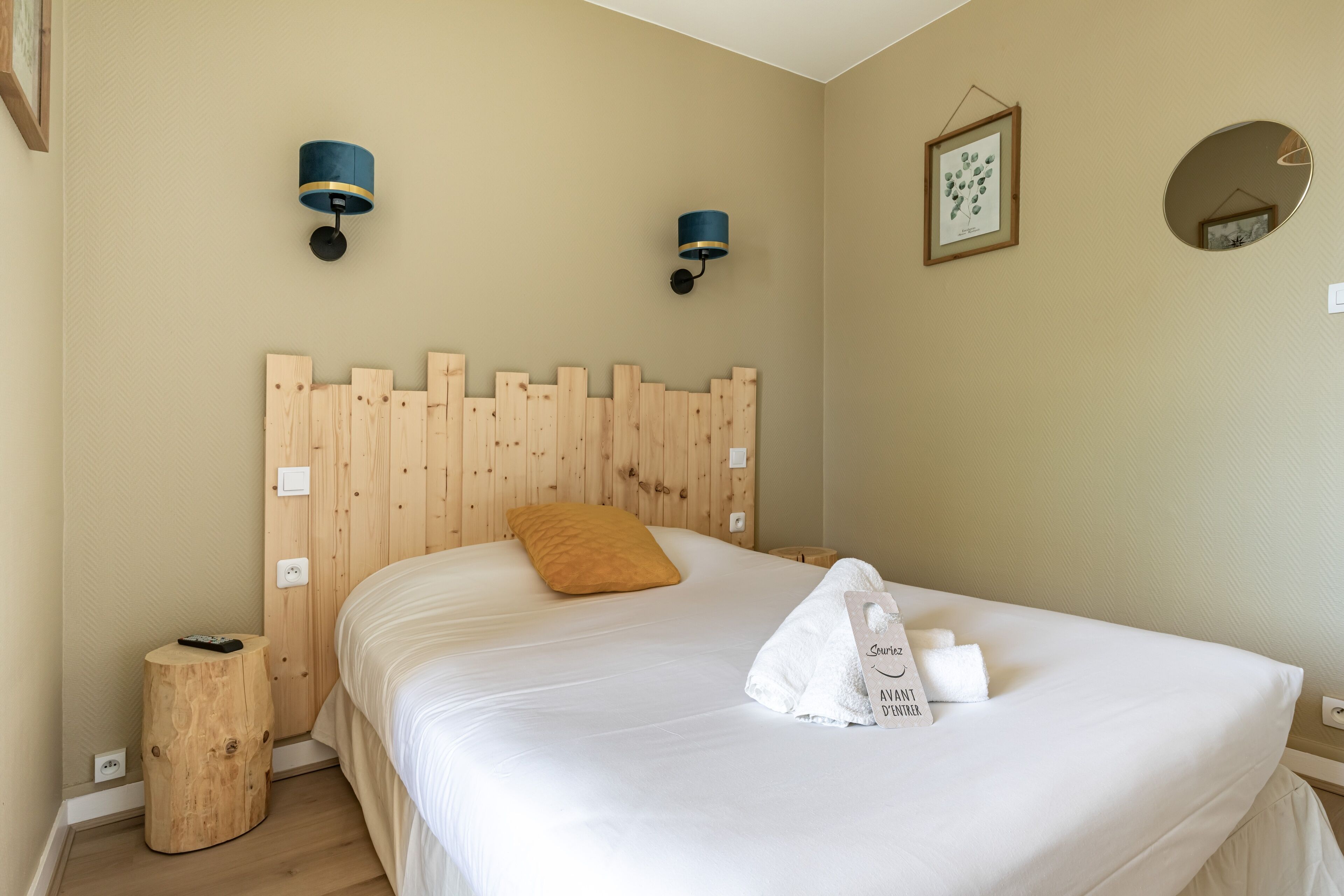 A neatly made bed with a wooden headboard, a small wooden stool, and a framed botanical print on the wall.